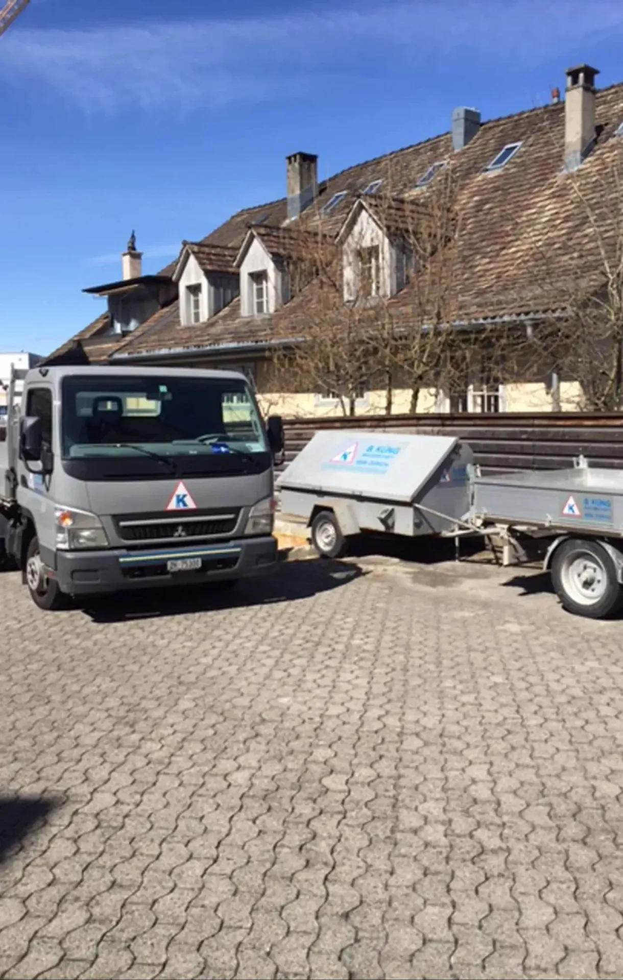 A grey Mitsubishi flatbed truck with Küng branding parked beside two company trailers on cobblestones.