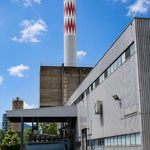 Industrial plant with a tall red-and-white chevron chimney stack alongside a large corrugated metal factory building.