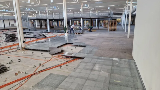 Workers lay modular floor tiles across a vast open hall with orange cable runs visible beneath.